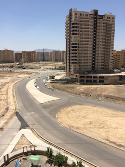 A tall residential building stands prominently against a bright blue sky. Surrounding it are several other apartment complexes in the background. The foreground features a newly constructed road and a roundabout with little traffic. The area appears to be under development, with bare soil patches and few green trees.