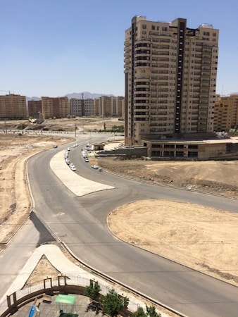 A tall residential building stands prominently against a bright blue sky. Surrounding it are several other apartment complexes in the background. The foreground features a newly constructed road and a roundabout with little traffic. The area appears to be under development, with bare soil patches and few green trees.