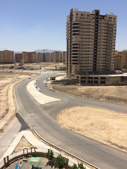 A tall residential building stands prominently against a bright blue sky. Surrounding it are several other apartment complexes in the background. The foreground features a newly constructed road and a roundabout with little traffic. The area appears to be under development, with bare soil patches and few green trees.