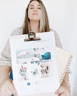 A person holding a clipboard with a mood board collage featuring images of a beach scene with an umbrella, a teal chair, succulents, and fruits. The mood board includes a palette of colors ranging from blue to light pastels. The person is also holding a decorative spiral-bound notebook with a gold pattern. The background is plain white.