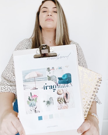 A person holding a clipboard with a mood board collage featuring images of a beach scene with an umbrella, a teal chair, succulents, and fruits. The mood board includes a palette of colors ranging from blue to light pastels. The person is also holding a decorative spiral-bound notebook with a gold pattern. The background is plain white.