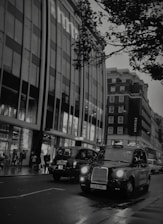 A street scene featuring classic black London taxis driving along a wet road, lined with modern buildings. The signage on the building indicates a well-known retail store, and several pedestrians are visible on the sidewalk, with tree branches hanging overhead.