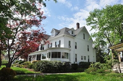 A large, white, two-story house with a gray roof surrounded by lush green trees and shrubs in a well-maintained garden. The house features classic architectural elements such as dormer windows and a bay window with a rounded design.