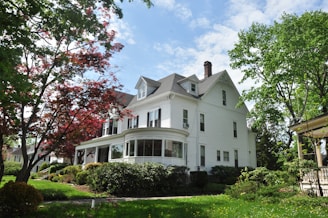 A large, white, two-story house with a gray roof surrounded by lush green trees and shrubs in a well-maintained garden. The house features classic architectural elements such as dormer windows and a bay window with a rounded design.