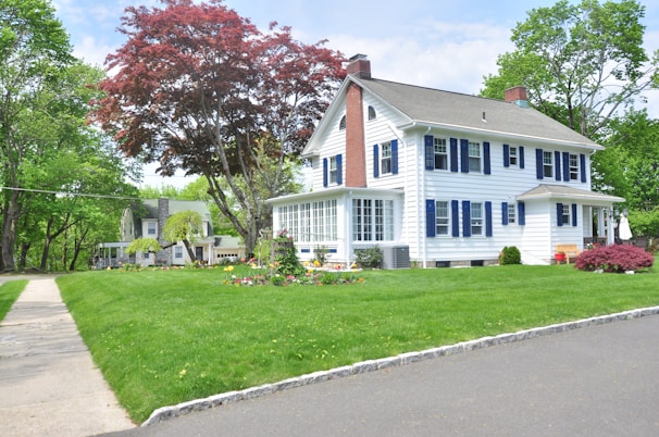A two-story house with white siding and blue shutters is set amid a lush, well-manicured lawn. There is a large tree with red leaves in the foreground and a variety of other trees and plants in the background. A sidewalk runs along the left side of the house, and a smaller house can be seen in the distance. The sky is partly cloudy.