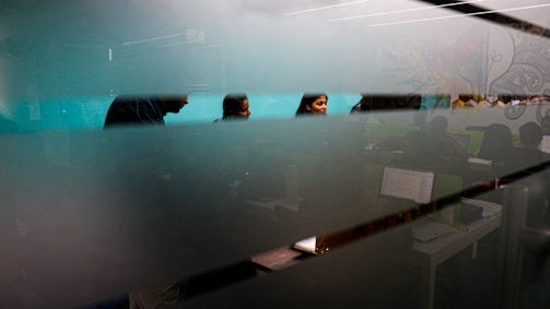 A group of people are visible through a frosted glass wall or partition. The setting appears to be an office or classroom, with individuals seated at desks working on computers. The image is partially obscured by horizontal bands of the frosted glass, and the room is dimly lit with a focus on the faces of the people in the background.