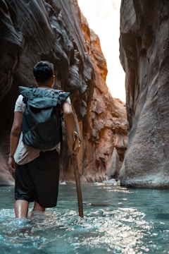 A person with a backpack wades through a narrow canyon filled with clear water, holding a wooden stick for support. The towering rock walls around them have a rough, textured surface and are illuminated by soft, natural light.
