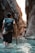 A person with a backpack wades through a narrow canyon filled with clear water, holding a wooden stick for support. The towering rock walls around them have a rough, textured surface and are illuminated by soft, natural light.