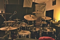 A dimly lit music studio featuring a drum set with multiple cymbals, drums, and a microphone. Acoustic panels and lighting equipment are in the background, creating a cozy and professional ambiance.