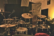 A dimly lit music studio featuring a drum set with multiple cymbals, drums, and a microphone. Acoustic panels and lighting equipment are in the background, creating a cozy and professional ambiance.