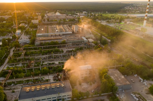 An aerial view of an industrial area at sunset with several large buildings and structures surrounded by greenery. Smoke or steam rises from parts of the complex, and the sunlight casts a warm glow over the scene. A tall smokestack is visible to the right, and residential areas are seen in the distance.