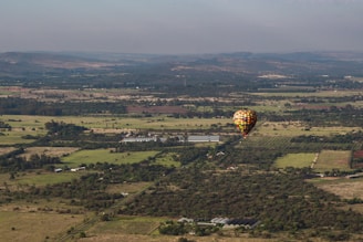 A hot air balloon floats above an expansive rural landscape. The balloon is colorful, standing out against fields and patches of trees. Distant hills and a vast sky are visible in the background, creating a feeling of openness and tranquility.