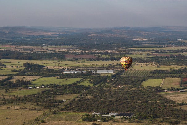 A hot air balloon floats above an expansive rural landscape. The balloon is colorful, standing out against fields and patches of trees. Distant hills and a vast sky are visible in the background, creating a feeling of openness and tranquility.