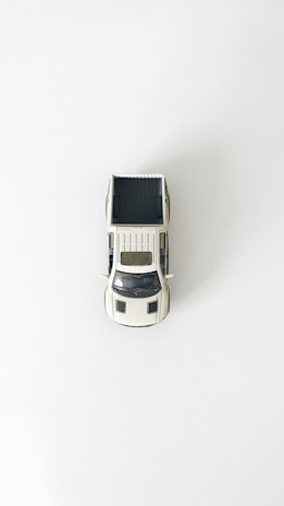 A miniature model of a white pickup truck is placed on a plain white background. The truck is viewed from a top-down perspective, highlighting its truck bed and the detailing on its roof and hood.