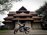 A traditional South Indian temple with intricate architectural details and terracotta-tiled roofs, set amidst a lush green environment with a classic motorcycle parked in front of it.