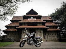 A traditional South Indian temple with intricate architectural details and terracotta-tiled roofs, set amidst a lush green environment with a classic motorcycle parked in front of it.