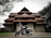A traditional South Indian temple with intricate architectural details and terracotta-tiled roofs, set amidst a lush green environment with a classic motorcycle parked in front of it.