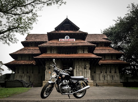A traditional South Indian temple with intricate architectural details and terracotta-tiled roofs, set amidst a lush green environment with a classic motorcycle parked in front of it.