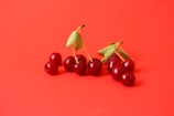 A small cluster of red cherries with green stems and leaves is arranged on a vibrant red background. The cherries are glossy and freshly picked, with some leaves still attached.