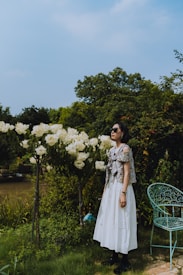 A woman in a floral top and white skirt stands in a garden filled with lush greenery and blooming white hydrangeas. She is wearing sunglasses and gazes upward, surrounded by a peaceful natural setting. An intricately designed green metal chair is nearby, adding to the serene atmosphere.