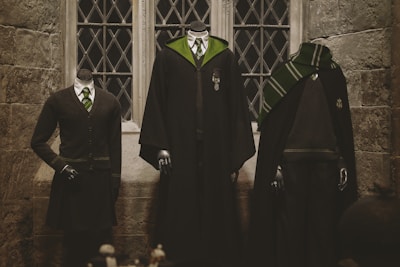 Three headless mannequins are dressed in outfits that resemble school uniforms typical of a fantasy setting. The central outfit is a long black robe with green lining and a silver emblem. To the left is a formal cardigan and tie ensemble with a skirt, and to the right is a caped outfit with a green and silver scarf. They are positioned against a stone wall with a multi-paned window in the background.