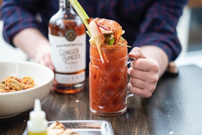 A person is holding a spicy Bloody Mary cocktail garnished with celery, a leafy green, and crab legs. In the background, there is a bottle labeled Ginger Spiced, likely a type of liquor. A bowl of food with a shellfish and sauce is positioned nearby on the dark wooden table.