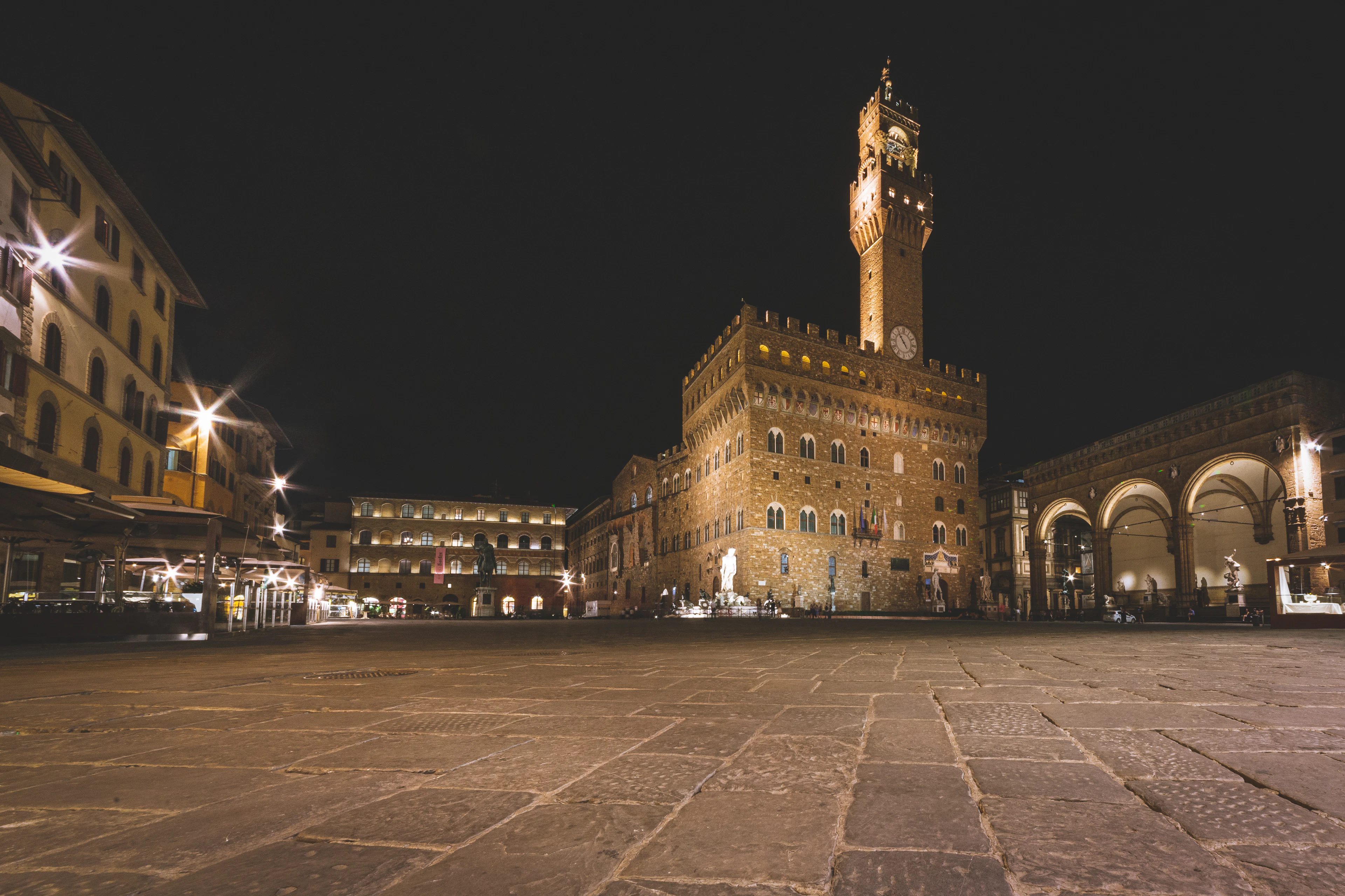 Piazza della Signoria in Florence at night, the floodlit Palazzo Vecchio and its crenellated tower standing over the empty stone square, with the lit arches of the Loggia dei Lanzi to the right.