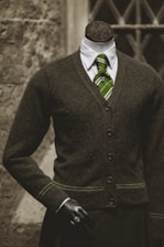 A headless mannequin dressed in a professional dark brown cardigan over a white shirt, complemented by a green striped tie. The mannequin has a black hand visible and stands in front of a textured stone wall with a window having crisscross metal bars.