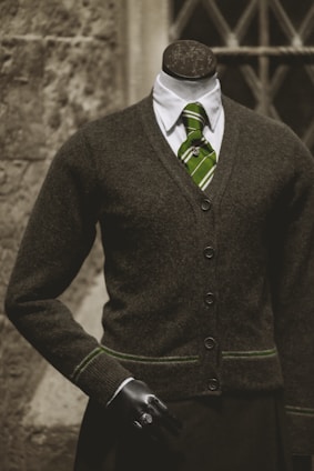 A headless mannequin dressed in a professional dark brown cardigan over a white shirt, complemented by a green striped tie. The mannequin has a black hand visible and stands in front of a textured stone wall with a window having crisscross metal bars.