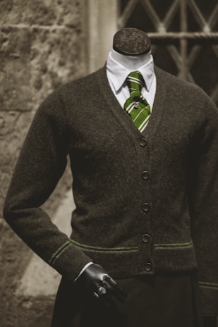 A headless mannequin dressed in a professional dark brown cardigan over a white shirt, complemented by a green striped tie. The mannequin has a black hand visible and stands in front of a textured stone wall with a window having crisscross metal bars.