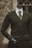 A headless mannequin dressed in a professional dark brown cardigan over a white shirt, complemented by a green striped tie. The mannequin has a black hand visible and stands in front of a textured stone wall with a window having crisscross metal bars.