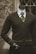 A headless mannequin dressed in a professional dark brown cardigan over a white shirt, complemented by a green striped tie. The mannequin has a black hand visible and stands in front of a textured stone wall with a window having crisscross metal bars.