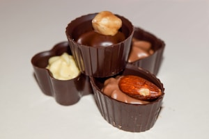 Three pieces of gourmet chocolate arranged on a light surface. Each piece is uniquely crafted with different types of nuts on top, including a hazelnut and an almond, and filled with different flavors. The chocolate is presented in a visually appealing manner with smooth textures and glossy surfaces.