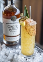 A tall cocktail glass filled with a crushed ice beverage, garnished with a slice of pineapple, a small pickle, and dusted with red spice. A bottle of ginger spiced whiskey is positioned in the background, slightly out of focus, on a surface covered with ice cubes.