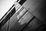 A black and white photograph of a modern building facade with an upward perspective. The structure features a series of vertically aligned panels that create geometric patterns. The contrast between light and shadow emphasizes the textures and lines, giving the image a dramatic and dynamic feel.