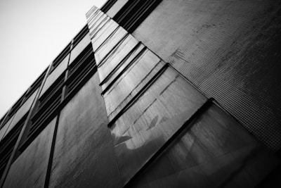 A black and white photograph of a modern building facade with an upward perspective. The structure features a series of vertically aligned panels that create geometric patterns. The contrast between light and shadow emphasizes the textures and lines, giving the image a dramatic and dynamic feel.