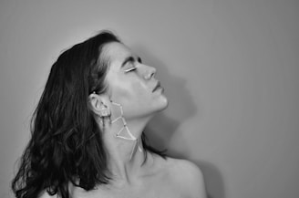 A black and white portrait of a person with medium-length dark hair gazing upward. The person has unique face and ear adornments that appear artistic.