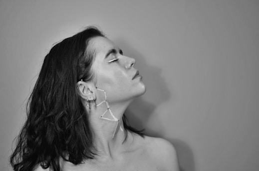 A black and white portrait of a person with medium-length dark hair gazing upward. The person has unique face and ear adornments that appear artistic.