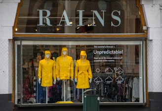 Three mannequins dressed in bright yellow raincoats are displayed in a shop window. Above the mannequins, large letters spell out 'RAINS' and to the side, there’s text reading 'Unpredictable!' suggesting a focus on waterproof essentials. The background of the window shows various clothing items.