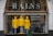Three mannequins dressed in bright yellow raincoats are displayed in a shop window. Above the mannequins, large letters spell out 'RAINS' and to the side, there’s text reading 'Unpredictable!' suggesting a focus on waterproof essentials. The background of the window shows various clothing items.