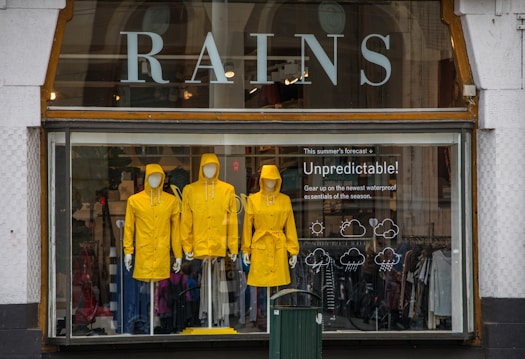 Three mannequins dressed in bright yellow raincoats are displayed in a shop window. Above the mannequins, large letters spell out 'RAINS' and to the side, there&rsquo;s text reading 'Unpredictable!' suggesting a focus on waterproof essentials. The background of the window shows various clothing items.