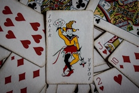 A collection of playing cards featuring a prominent Joker card in the center. Surrounding it are various cards from a deck, including hearts and diamonds, some with intricate designs and patterns. The Joker is depicted in a colorful outfit holding a set of cards.