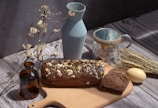 A rustic wooden table setting featuring a loaf of dark bread topped with nuts and seeds, partially sliced on a cutting board. Surrounding it are a light blue ceramic vase, a brown glass bottle, a metal mug labeled 'Home & Flower', some wheat stalks, an egg, and decorative dry branches.