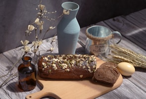A rustic wooden table setting featuring a loaf of dark bread topped with nuts and seeds, partially sliced on a cutting board. Surrounding it are a light blue ceramic vase, a brown glass bottle, a metal mug labeled 'Home & Flower', some wheat stalks, an egg, and decorative dry branches.