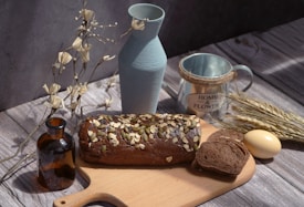 A rustic wooden table setting featuring a loaf of dark bread topped with nuts and seeds, partially sliced on a cutting board. Surrounding it are a light blue ceramic vase, a brown glass bottle, a metal mug labeled 'Home & Flower', some wheat stalks, an egg, and decorative dry branches.
