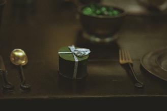 A small, heart-shaped, dark green gift box with a silver ribbon is placed on a dark surface. It is surrounded by a golden spoon on the left and a dark fork on the right. In the background, a bowl filled with shiny green objects adds a subtle highlight to the dimly lit scene.