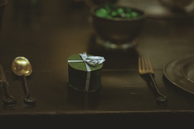 A small, heart-shaped, dark green gift box with a silver ribbon is placed on a dark surface. It is surrounded by a golden spoon on the left and a dark fork on the right. In the background, a bowl filled with shiny green objects adds a subtle highlight to the dimly lit scene.