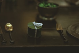 A small, heart-shaped, dark green gift box with a silver ribbon is placed on a dark surface. It is surrounded by a golden spoon on the left and a dark fork on the right. In the background, a bowl filled with shiny green objects adds a subtle highlight to the dimly lit scene.