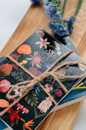 A notebook with a dark cover adorned with various colorful floral illustrations is tied with a rustic twine. It rests on a wooden surface, next to some vibrant blue flowers.
