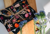 A collection of floral-themed postcards is tied together with rustic twine, resting on a wooden surface. Each postcard features vibrant, colorful illustrations of various flowers. In the foreground, there is a sprig of purple flowers casting a soft shadow.